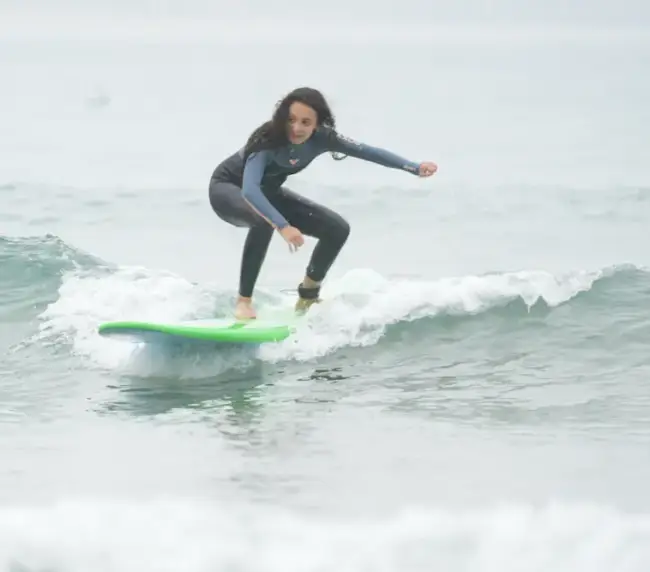 Woman in a black wetsuit riding a small wave on a green surfboard during a beginner surf session in Imsouane.