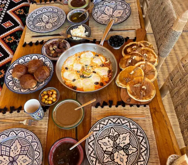 Breakfast table with eggs, pancakes, croissants, dates, bread, and juice at an Imsouane surf camp.