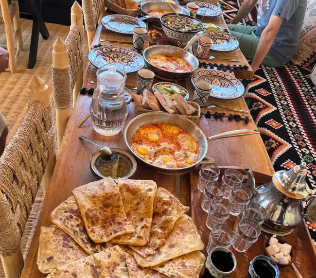 Guest seated at a Moroccan breakfast table with flatbread, eggs, salads, and mint tea in an Imsouane surf camp.