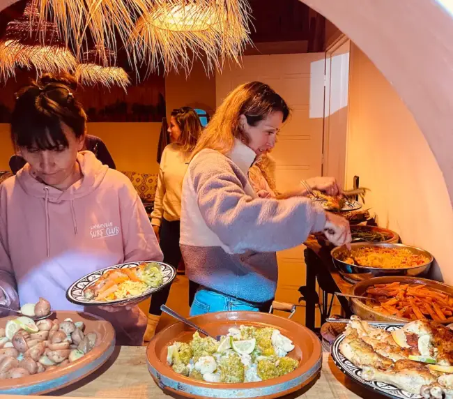 Surf house guests serving vegetables, rice, and grilled fish from a shared dinner buffet in Imsouane.