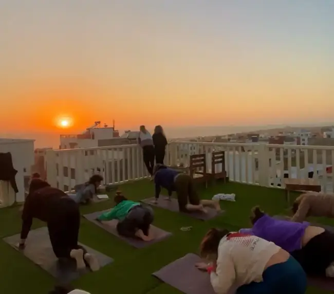Surf house guests practicing yoga on rooftop mats as the sun sets over Imsouane and the Atlantic.