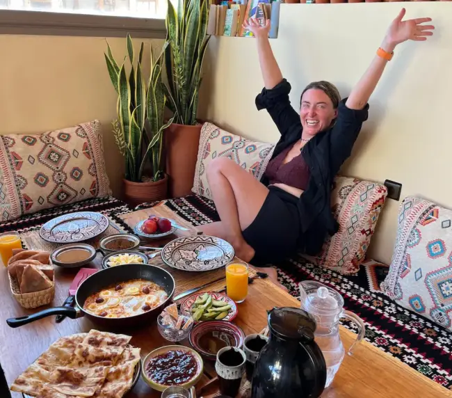 Smiling guest raises both arms behind a Moroccan breakfast spread at an Imsouane surf house.
